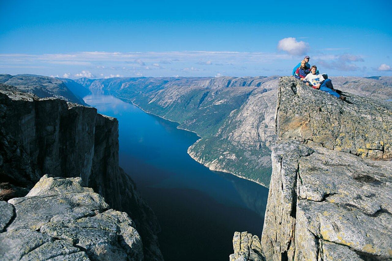 Looking straight down a cliff into the deep blue Lysefjorden, with three hikers sitting on the rock edge