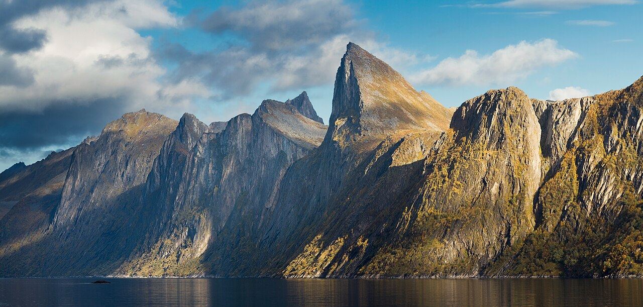 The sail-shaped peak of Segla rising straight out of a calm, dark fjord in warm autumn light